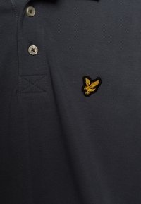 Polo de color gris oscuro hecho de tela texturada, con un pequeño logo bordado de un águila negra y dorada en el pecho y detalles de botones en el cuello.
