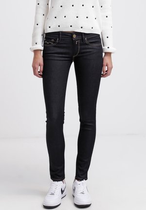 Person wearing black skinny jeans, white sneakers with black swoosh, and a white sweater with black polka dots, standing against plain background.