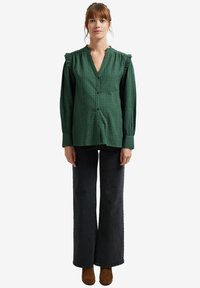 Green button-up shirt with ruffled shoulders, textured finish, long sleeves, and black wide-leg jeans. Brown heeled footwear completes the look.
