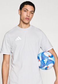 Λευκό κοντομάνικο μπλουζάκι με κεντημένο λογότυπο τριών ρίγων. Μπλε ποδοσφαιρική μπάλα με κόκκινες και λευκές λεπτομέρειες κρατιέται κοντά στο σώμα.