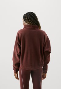 Burgunderfarbener Fleece-Pullover mit hohem Kragen, entspannter Passform und überschnittenen Schultern. Texturiertes Material; kombiniert mit passender Leggings.