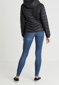 Doudoune noire avec capuche, arborant un motif matelassé en chevrons. Portée avec un jean skinny bleu et des sneakers noires à motifs.