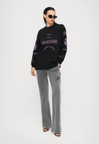 Naine, kes kannab musta ülemäära suurt sweatshirt'i lilla tekstiga, halli laiade jalgade pükse, päikeseglasse ja kontsasaapaid, seismas valge tausta ees.