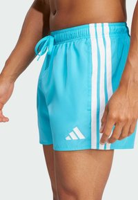 Svetlomodré plavky vyrobené z hladkej tkaniny, s elastickým pásom na šnúrku a tromi bielymi pruhmi po stranách.