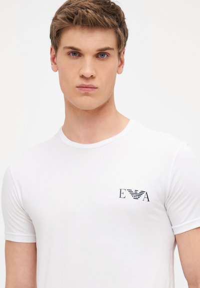 White cotton t-shirt with short sleeves, crew neck, and a small black logo featuring "EVA" on the left chest area.