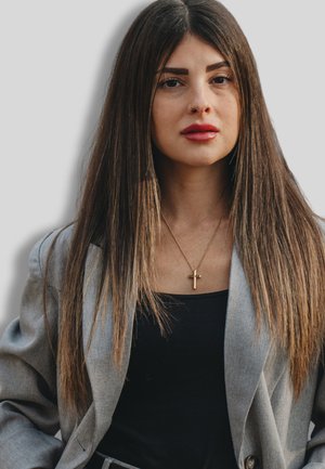 Frau mit langen, glatten braunen Haaren, die ein graues Blazer über einem schwarzen Oberteil und eine goldene Kreuzkette trägt. Schlicher Hintergrund.