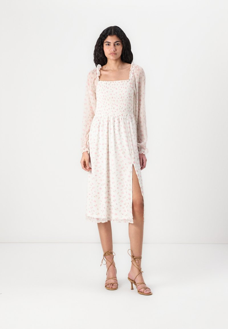 Rochie ușoară, cu imprimeu floral, cu un corp strâns, mâneci lungi transparente, fante laterale și tiv în formă de val. Sandale cu curele completează aspectul.