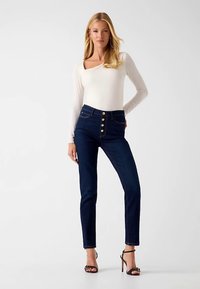 Donkerblauwe jeans met hoge taille, vijf knopen aan de voorkant, verticale stikaccenten en een slanke pasvorm, gecombineerd met een aangesloten witte top met lange mouwen.