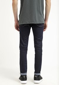 Vaqueros ajustados de color azul oscuro con un sutil desgaste, dobladillos remangados y costuras naranjas, combinados con zapatillas negras con suelas blancas.