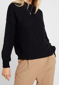 Black knitted sweater with a ribbed texture, long sleeves, and a relaxed fit. Paired with beige trousers. Sleek and minimal design.