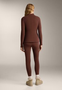 Smeđi prilagođeni dugoročni rukavi i tajice, izrađeni od glatkog materijala. Model nosi svijetle, plišane čarape i bež gležnjače.