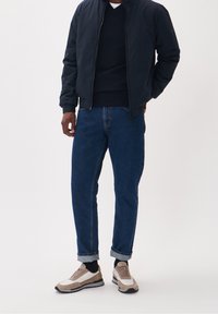 Giacca bombardiere navy, maglione scuro, jeans blu con maniche arrotolate e sneakers bicolore con accenti beige e pannelli bianchi. Design semplice, vestibilità casual.