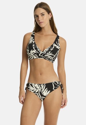 Kobieta o długich brązowych włosach, ubrana w czarno-biały bikini w tropikalny wzór, z szerokimi ramiączkami i majtkami z wiązaniem po bokach, stojąca na białym tle.