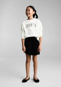 Bela sweatshirt z okrasjem "JOYFUL", v kombinaciji z črno teksturirano krilo z metuljčkom in pikčastim vzorcem ter črnimi balerinkami.