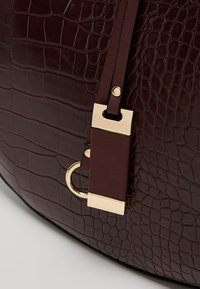 Viininpunainen krokotiilikuvioitu käsilaukku, jossa on kullanväriset metalliosat ja litteä, säädettävä hihna. Tasainen, teksturoitu pinta.