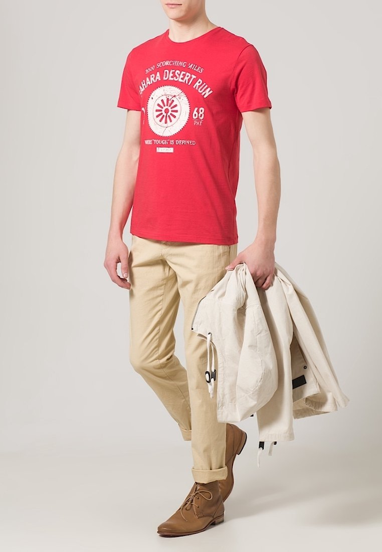 Rotes Baumwoll-T-Shirt mit weißem Text und Grafik, beige Chinos, braune schnürbare Stiefel und eine hellfarbige Jacke über dem Arm getragen.