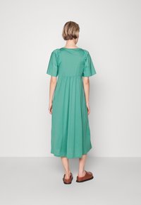 Robe teal jusqu'aux genoux avec des manches courtes en forme de poire, une taille froncée et une texture lisse. Associée à des chaussures marron à enfiler.