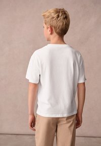 Pojke med kort blont hår står med ryggen mot kameran och bär en enfärgad vit t-shirt med korta ärmar och beige byxor mot en neutral bakgrund.