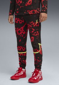 A person wears black pants with a red and yellow abstract design, complemented by bright red sneakers, against a gradient background.