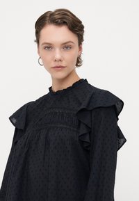 Blusa nera realizzata in un tessuto con texture, con maniche voluminose e un collo alto, caratterizzata da delicati bordi in pizzo e piccoli punti in rilievo.