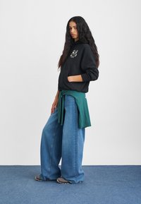 Fekete kapucnis pulóver grafikus mintával, bőszárú kék farmerrel és derékban megkötött zöld pólóval párosítva. A modell szürke cipőt visel.