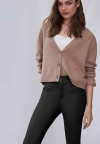 Cárdigan rosa claro hecho de material suave, con escote en V, mangas largas y cierre de botones, combinado con jeans ajustados negros.