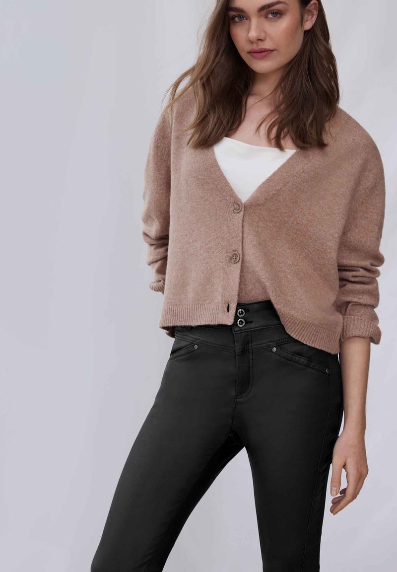 Cárdigan rosa claro hecho de material suave, con escote en V, mangas largas y cierre de botones, combinado con jeans ajustados negros.