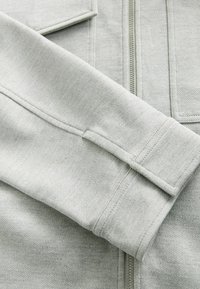 Light gray jacket featuring a textured fabric, zipper closure, and folded sleeves with stitched accents and patch pockets.