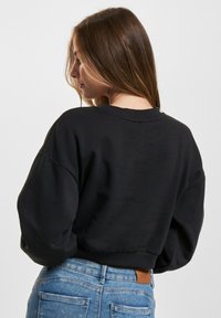 Svart kort sweatshirt med rund hals og senkede skuldre, med en jevn tekstur og ribbede mansjetter, iført sammen med blå jeans.