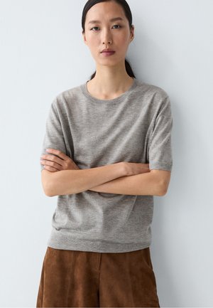Donna in piedi con le braccia incrociate che indossa un maglione grigio a maniche corte e una gonna marrone su uno sfondo bianco uniforme.