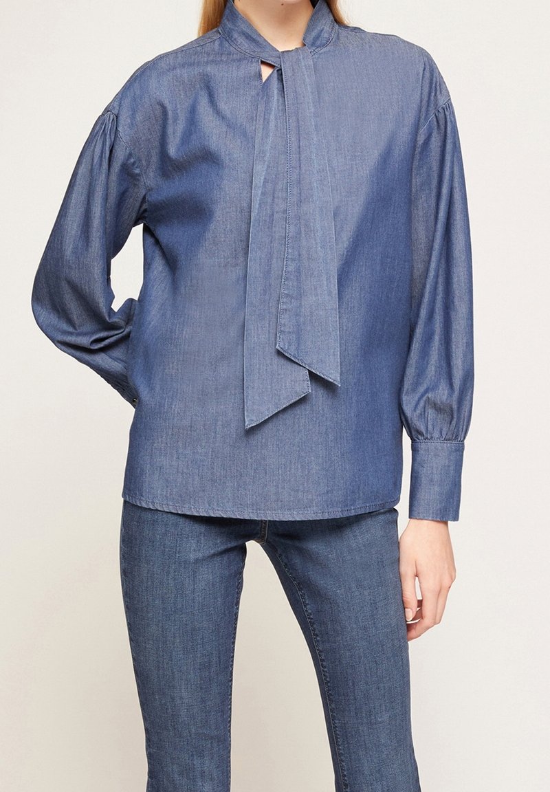Femme portant un chemisier en jean bleu avec des manches longues bouffantes et un détail noué au col, associé à un jean skinny bleu.
