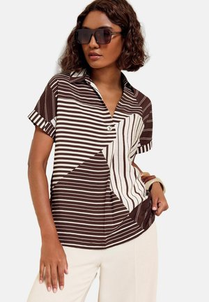 Woman wearing oversized brown and white striped short-sleeve shirt, light pants, large square sunglasses, and brown and white bracelets.