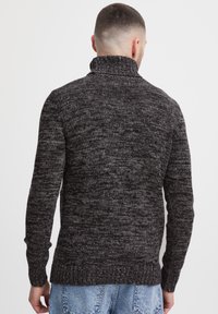 Man som bär en mörkgrå melerad stickad polotröja och blå jeans, sedd bakifrån mot en vit bakgrund.