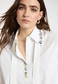 Joven con cabello castaño suelto que lleva una camiseta blanca con adornos de gemas multicolores en el cuello y la tapeta.