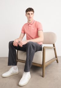 Polo shirt rose clair avec col et poignets contrastants, porté avec un jean gris foncé et des baskets blanches, assis sur une chaise en bois minimaliste.
