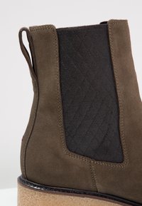 Botte Chelsea en daim marron avec panneau latéral élastique matelassé noir et semelle en crêpe beige, montrant des coutures détaillées et une languette de tirage à l'arrière.