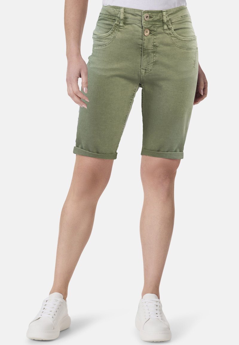 Grüne Denim-Bermudashorts mit glatter Textur, fünf Taschen, umgeschlagenen Säumen und zwei Knopfverschlüssen vorne. Kombiniert mit weißen Schuhen.