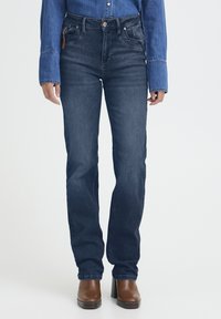 Donna che indossa jeans blu scuro a gamba dritta, stivali con tacco marroni e una camicia di jeans a maniche lunghe su uno sfondo bianco uniforme.