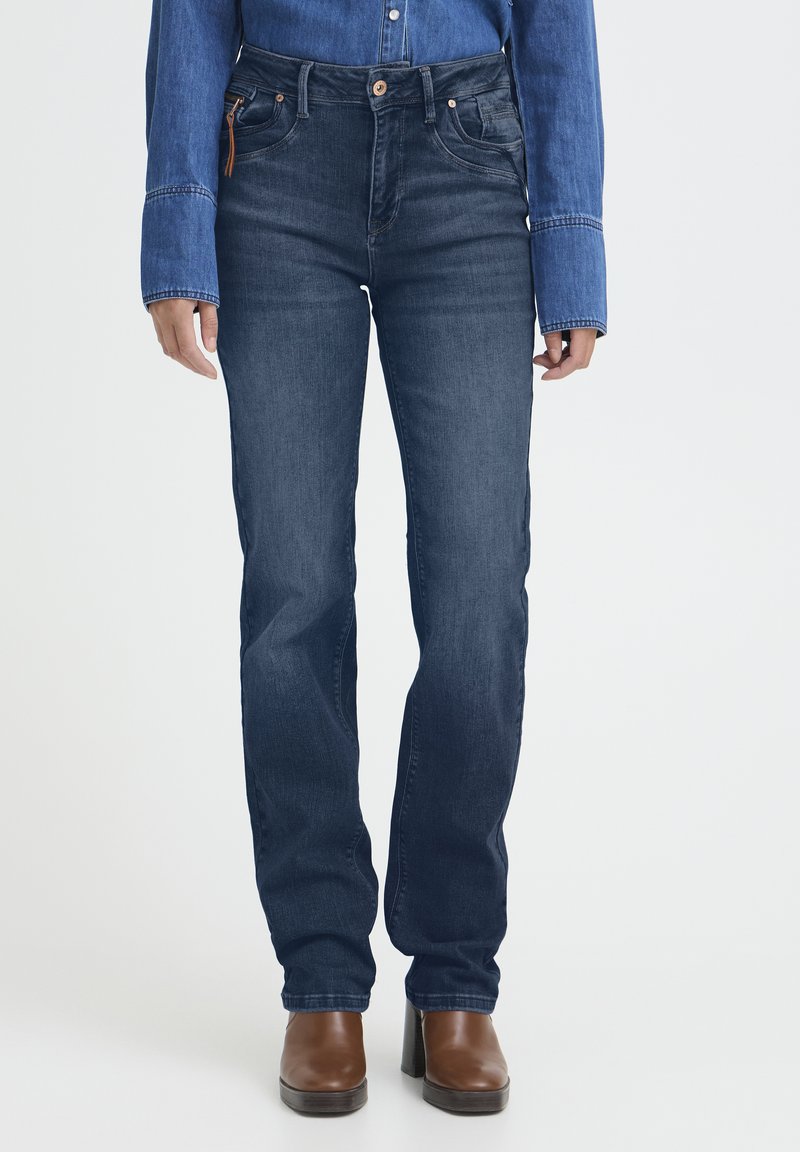 Donna che indossa jeans blu scuro a gamba dritta, stivali con tacco marroni e una camicia di jeans a maniche lunghe su uno sfondo bianco uniforme.