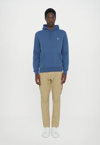 Mėlynas medvilninis своими s dugnu kišenėmis ir sustringa gobtuvu, derinamas su smėlio spalvos kargo kelnėmis ir baltais sportbačiais, stovintis ant balto fono.