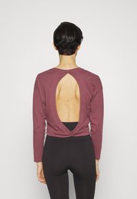 Persona con capelli corti e scuri che indossa una maglietta bordeaux a maniche lunghe con schiena ovale aperta e pantaloni aderenti neri, in piedi davanti a uno sfondo semplice.