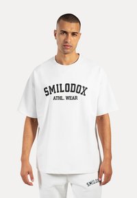 Weißes Baumwoll-T-Shirt mit Rundhalsausschnitt und überschnittenen Schultern, mit "SMILODOX ATHL. WEAR" in fettem Schwarz auf der Vorderseite bedruckt.