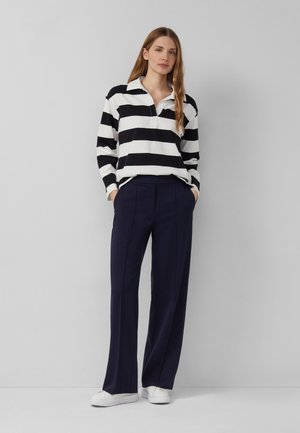 Femme debout contre un mur gris, portant un chemisier à rayures noires et blanches à manches longues, un pantalon large bleu marine et des baskets blanches.