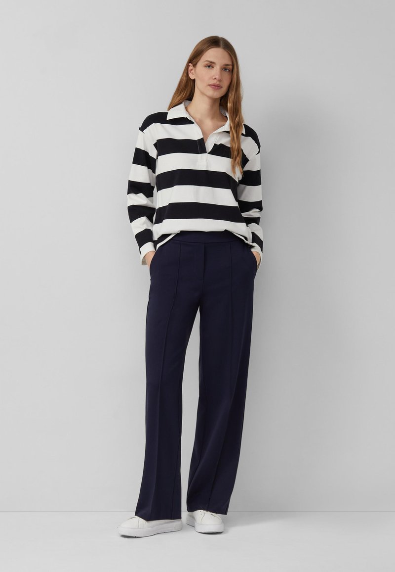 Žena stojaca opretá o sivú stenu, oblečená v čierno-bielom pruhovanom tričku s dlhým rukávom, tmavomodrých nohaviciach s širokými nohavicami a bielych teniskách.