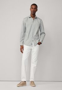 Camicia a righe blu chiaro e bianca, abbinata a pantaloni bianchi. Il modello è in piedi con le mani in tasca, indossando scarpe beige.
