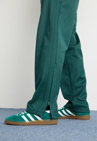 Vihreät urheiluhousut, joissa on sivuvetoketjut, yhdistettyinä vihreisiin mokkakenkiin, joissa on valkoiset raidat ja kumipohja. Matto lattian alla.