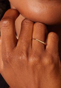 Gouden ring op een hand met gestructureerde bruine huid. De ring heeft een dunne band en een glad, rechthoekig bovenoppervlak.