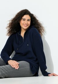 Mörkblå huvtröja med ribbade ärmar, halvdragkedja fram och mjuk stickad textur. Matchad med grå byxor.