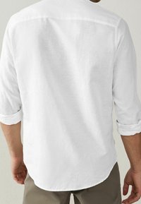 Bílá lněná košile s ohrnutými rukávy a zakulaceným lemem; texturovaná tkanina poskytuje lehký a prodyšný vzhled zezadu.