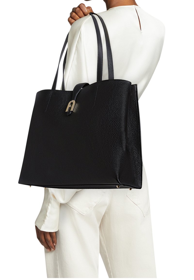 Personne portant une tenue blanche tenant un grand sac cabas en cuir noir texturé avec doubles poignées et fermoir métallique sur l'épaule.